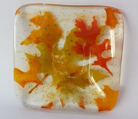 Small square fused glass dish with translucent autumn maple leaves in orange, red, and gold on a clear base with gold frit accents.